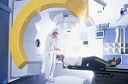 Titel: Fotograf / Plattform Patient liegt auf einer Liege, davor steht der Arzt, markant links im Bild ein golder Bogen des NovalisMRT-Geräts
